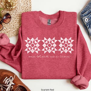 May include: Scarlett red sweatshirt with a white geometric design and the text "one block at a time." The long-sleeved crew neck top is made of a soft material.