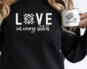 May include: Black sweatshirt with white text that reads "LOVE in every stitch". The text includes a white patchwork quilt block design in the center of the word "LOVE". A white enamel mug with a black outline of a spool of thread and needle is visible in the background.