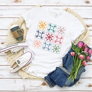 May include: A white t-shirt with a patchwork quilt design featuring nine colorful squares with a star pattern in each square. The squares are arranged in a 3x3 grid.