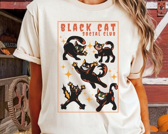 May include: Cream-colored t-shirt featuring a graphic of several black cats in various poses with green eyes and orange accents. The text "BLACK CAT SOCIAL CLUB" is displayed in an orange rectangular frame above the cats. The shirt has short sleeves.