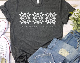 May include: Dark heather gray t-shirt with a white design of three quilt blocks and the text "one block at a time".