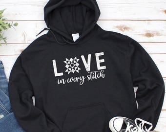 May include: Black hooded sweatshirt with the text "LOVE in every stitch" in white. The word "LOVE" features a quilt block design. The sweatshirt has a front pocket and a drawstring hood. A pair of black and white sneakers are visible.