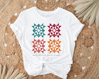 May include: White t-shirt with a design of four colorful quilt blocks. The text "one block at a time" is printed below the blocks.