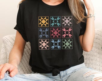May include: A black short-sleeved t-shirt featuring a graphic print of nine square quilt blocks arranged in a three-by-three grid. Each square contains a geometric star pattern in various muted colors including yellow, teal, mauve, blue, and pink.