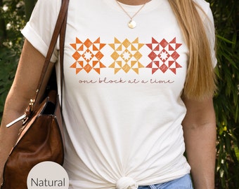 May include: Cream-colored t-shirt featuring three geometric quilt block designs in shades of orange and red. The text "one block at a time" is printed below the blocks in a handwritten style font. The shirt is a casual, everyday wear item.