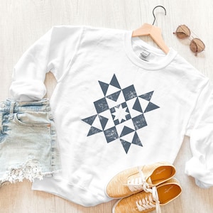 May include: White sweatshirt with a blue and white geometric pattern in the shape of a star. The sweatshirt is on a wooden surface with a pair of jeans and shoes.