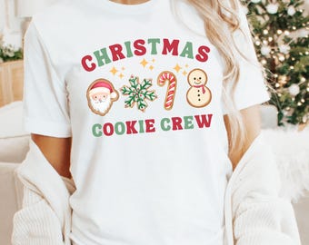 May include: White t-shirt with a festive Christmas design. The design includes the words "CHRISTMAS COOKIE CREW" in red and green, along with gingerbread cookie illustrations of Santa, a snowflake, a candy cane, and a snowman.