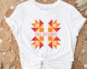 May include: White t-shirt featuring a colorful geometric design with the words "FALL BLESSINGS" in pink. The design incorporates red, orange, and yellow shapes, creating a fall-themed aesthetic. The shirt is made of a soft material.