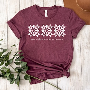 May include: A maroon t-shirt with a white design of three quilt blocks and the text "one block at a time".