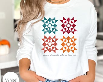 May include: White long-sleeved sweatshirt with a graphic of four colorful quilt blocks and the text "one block at a time".