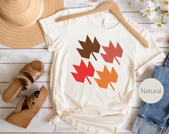 May include: Cream-colored t-shirt with a leaf design in shades of brown, red, and orange. The shirt is hanging on a wooden hanger. A straw hat and brown sandals are next to the shirt. The word "Natural" is printed on a white circle.