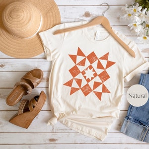May include: Cream-colored t-shirt with a geometric starburst design in shades of orange. The shirt is hanging on a wooden hanger. A straw hat and brown sandals are visible in the background. The word "Natural" is printed on a round label.