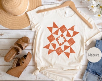 May include: Cream-colored t-shirt with a geometric starburst design in shades of orange. The shirt is hanging on a wooden hanger. A straw hat and brown sandals are visible in the background. The word "Natural" is printed on a round label.