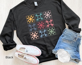 May include: Black sweatshirt with a colorful patchwork quilt design. The sweatshirt is shown laid flat on a wooden surface with a pair of white sneakers and denim shorts.