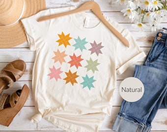 May include: A white t-shirt with a geometric pattern of colorful stars. The shirt is on a hanger and has the word "Natural" written on a tag.