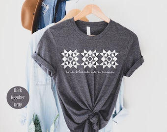 Op de afbeelding: Donker grijs t-shirt met een witte afbeelding van drie quiltblokken en de tekst "one block at a time".