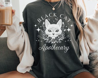 May include: Dark gray sweatshirt with a white graphic of a cat's face surrounded by flowers, stars, and the words "Black Cat Apothecary". The sweatshirt is a relaxed fit.