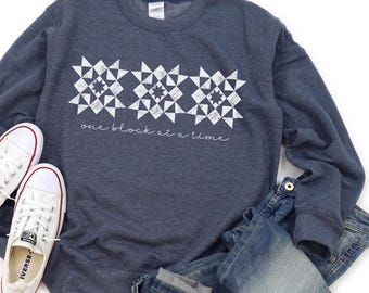 May include: A navy blue sweatshirt with a white geometric design and the text "one block at a time." White canvas sneakers and blue jeans are also in the image. The sweatshirt is a casual style.