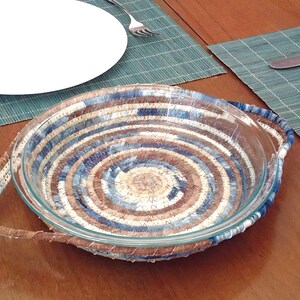 May include: A round glass baking dish with a woven denim-style fabric wrap. The fabric features shades of blue, brown, and cream in a spiral pattern. The dish has two handles and sits on a wooden table. A white plate and silverware are in the background.