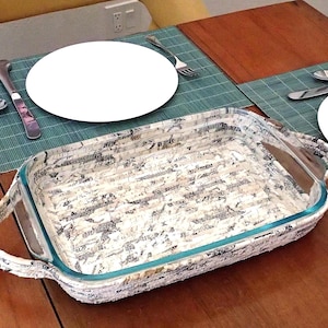 May include: A rectangular glass baking dish with a decorative woven paper exterior, featuring a black and white pattern. The dish has clear glass sides and handles. Set on a wooden table with place settings.