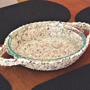 May include: A round glass pie dish with a woven, textured border and handles. The border has a light beige and brown pattern. The dish sits on a wooden surface, with black placemats in the background.