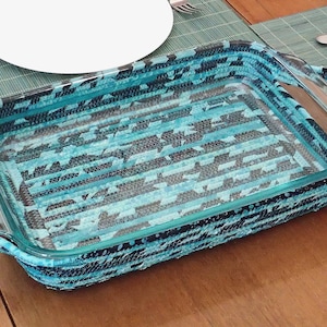 May include: A rectangular glass baking dish with a woven blue and black patterned exterior. The dish has two handles and is set on a wooden table with place settings.