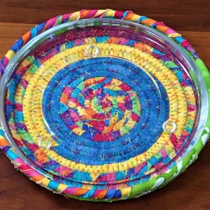 May include: A colorful fabric coaster with a glass insert. The coaster is made of strips of fabric sewn together in a spiral pattern. The fabric is in shades of blue, yellow, green, red, and pink. The glass insert is clear and has a smooth surface.