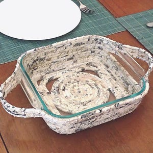 May include: A clear glass baking dish with a woven paper basket. The basket is made of cream-colored paper with black text and designs. The dish has a spiral design in the center and handles on each side.