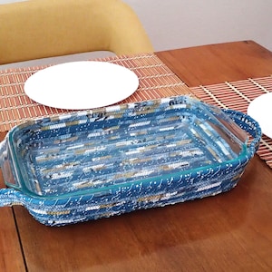 May include: A rectangular glass baking dish with woven blue, white, and gold fabric around the sides and handles. The dish sits on a wooden table with two white plates and bamboo placemats. The background includes a yellow chair.