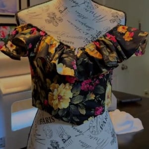 Könnte beinhalten: Ein schwarzes schulterfreies bauchfreies Oberteil mit einem lebendigen Blumenmuster aus gelben und rosa Blüten sowie grünen Blättern. Das Kleidungsstück hat einen gerüschten Ausschnitt und einen spitzen Saum, präsentiert auf einer Schneiderpuppe.
