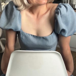Puede incluir: Una blusa azul claro con mangas abullonadas y cuello cuadrado. La blusa está hecha de una tela suave y se combina con unos vaqueros azules. La persona está sentada en una silla blanca en una cocina.