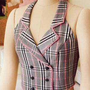 Pu&ograve; includere: Un gilet a quadri bianco e nero con bordi rosa e bottoni neri. Il gilet ha una scollatura halter e una chiusura a doppio petto.