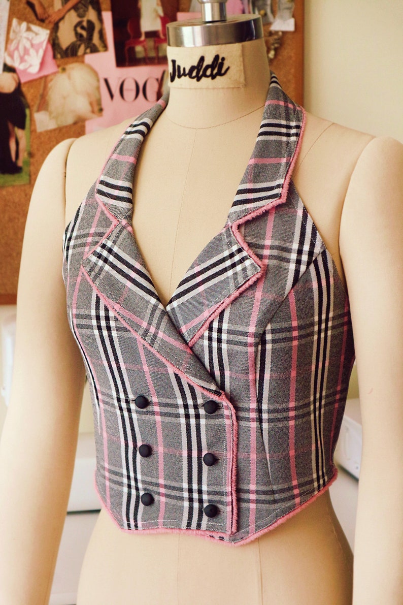 Pu&ograve; includere: Un gilet a quadri bianco e nero con bordi rosa e bottoni neri. Il gilet ha una scollatura halter e una chiusura a doppio petto.