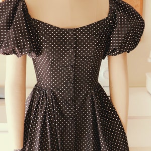 Puede incluir: Un vestido negro con lunares blancos y un frente abotonado. El vestido tiene mangas abullonadas y un corpi&ntilde;o ajustado.
