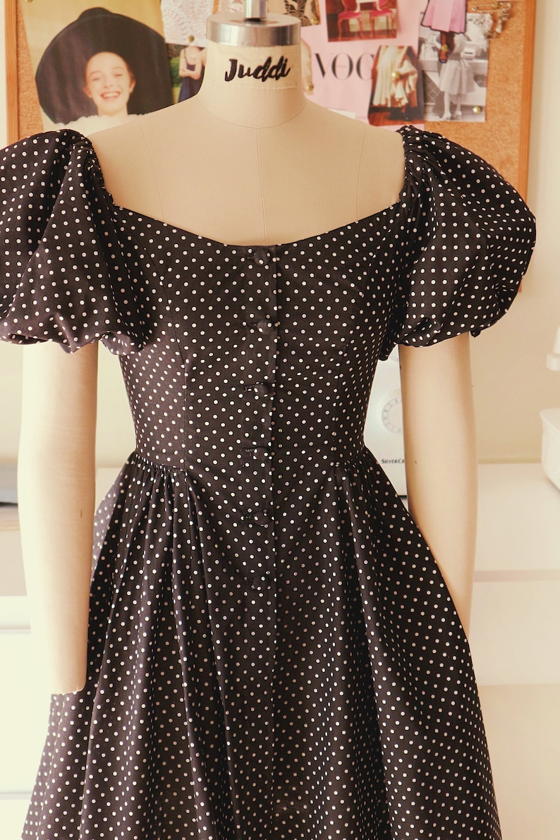 Puede incluir: Un vestido negro con lunares blancos y un frente abotonado. El vestido tiene mangas abullonadas y un corpi&ntilde;o ajustado.