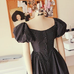 Puede incluir: Un vestido de lunares negros y blancos con mangas abullonadas y escote de coraz&oacute;n. El vestido est&aacute; en un maniqu&iacute; con el nombre "Juddi" en el busto.