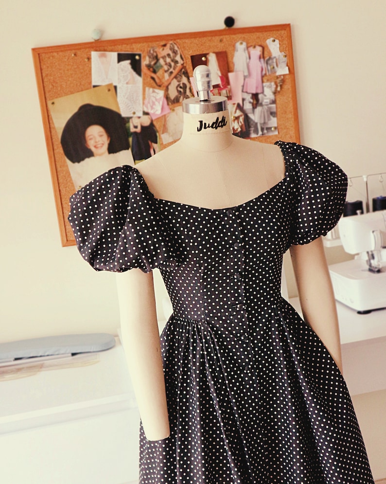 Puede incluir: Un vestido de lunares negros y blancos con mangas abullonadas y escote de coraz&oacute;n. El vestido est&aacute; en un maniqu&iacute; con el nombre "Juddi" en el busto.