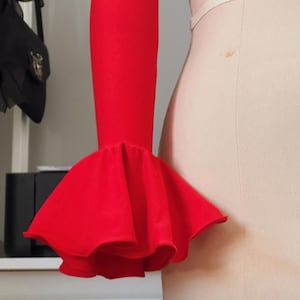 May include: A bright red sleeve with a flared ruffle detail. The sleeve is attached to a mannequin, highlighting the design and fabric. The ruffle adds a dramatic flair to the garment.
