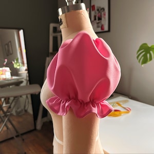 May include: A pink fabric puff sleeve with a ruffled edge. The sleeve is attached to a dress form.