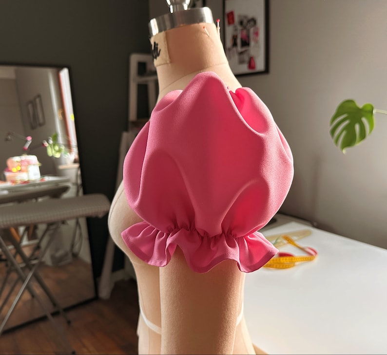 May include: A pink fabric puff sleeve with a ruffled edge. The sleeve is attached to a dress form.