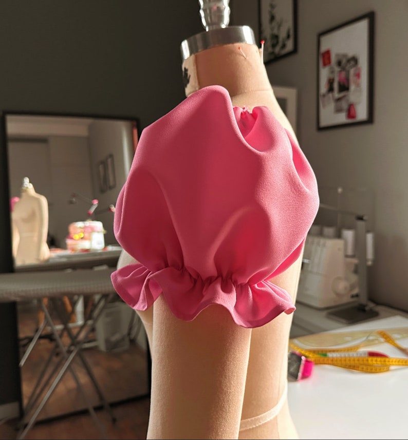 May include: A pink fabric puff sleeve with a ruffled edge. The sleeve is on a dress form.