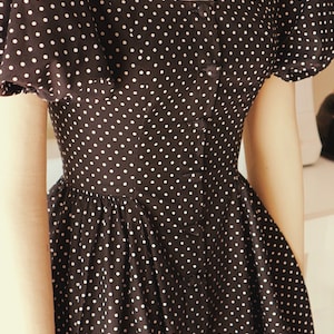 Puede incluir: Un vestido negro con lunares blancos y un frente abotonado. El vestido tiene mangas abullonadas y un corpi&ntilde;o ajustado.