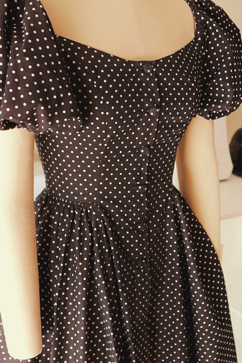 Puede incluir: Un vestido negro con lunares blancos y un frente abotonado. El vestido tiene mangas abullonadas y un corpi&ntilde;o ajustado.