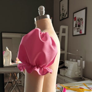 May include: A pink fabric puff sleeve on a dress form. The sleeve has a ruffled edge.
