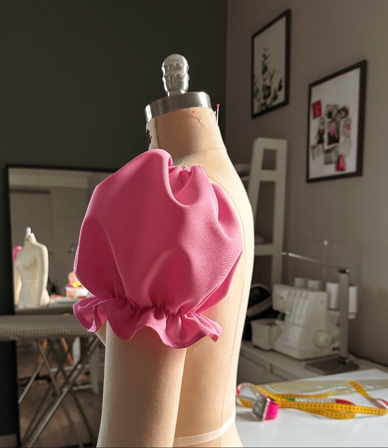 May include: A pink fabric puff sleeve on a dress form. The sleeve has a ruffled edge.