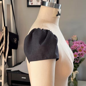 May include: A close-up of a dark grey fabric sleeve on a dress form. The sleeve has a gathered, puffed look at the shoulder.