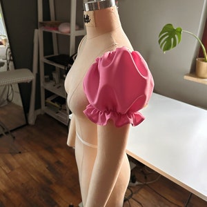 May include: A pink fabric puff sleeve with a ruffled edge, attached to a dress form.