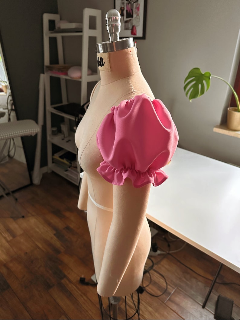May include: A pink fabric puff sleeve with a ruffled edge, attached to a dress form.