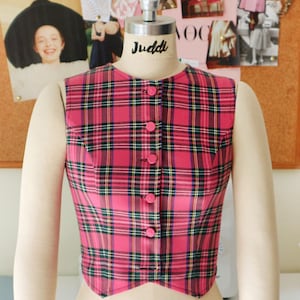 May include: A pink and black plaid sleeveless waistcoat with a pointed hem and button fastening. The waistcoat is on a mannequin.