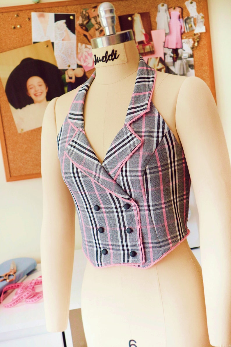 Pu&ograve; includere: Un top halter a quadri grigi e rosa con chiusura a doppio petto e bottoni neri. Il top ha un bordo sfrangiato ed &egrave; su un manichino.